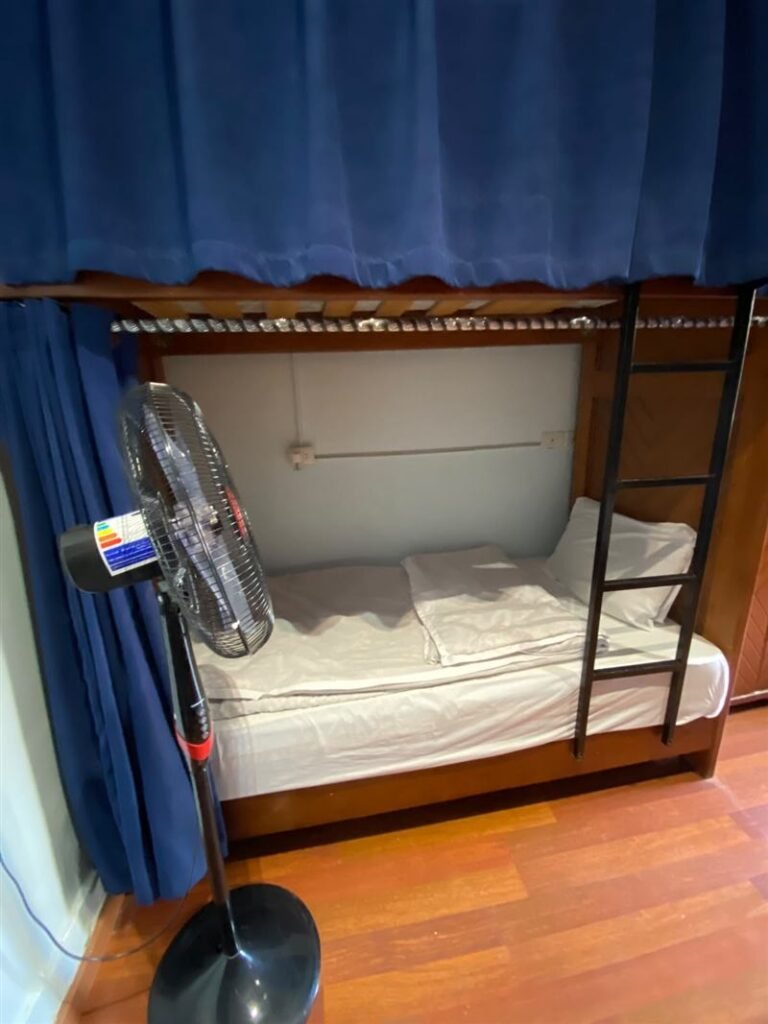 Lower bunk bed with a blue curtain and a floor fan in the dorm